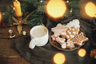 Christmas gingerbread cookies, coffee in stylish white cup, fir branches, warm lights on napkin and rustic wooden table. Atmospheric image. Winter countryside hygge. Happy Holidays!
