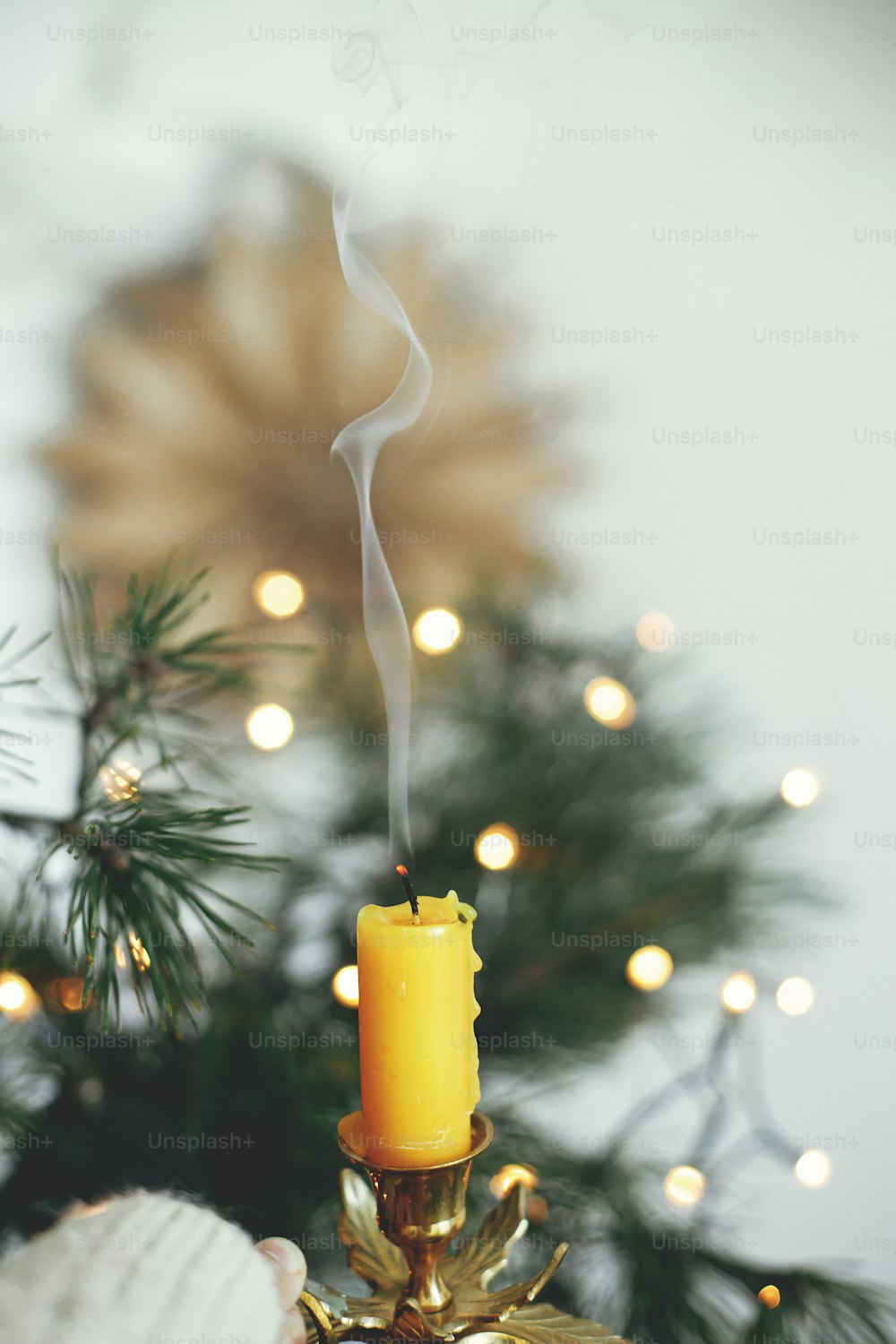 Hand holding stylish vintage candlestick with extinguish candle and smoke on background of warm lights, fir branches and sweden star in festive scandinavian room. Atmospheric hygge home. Relax
