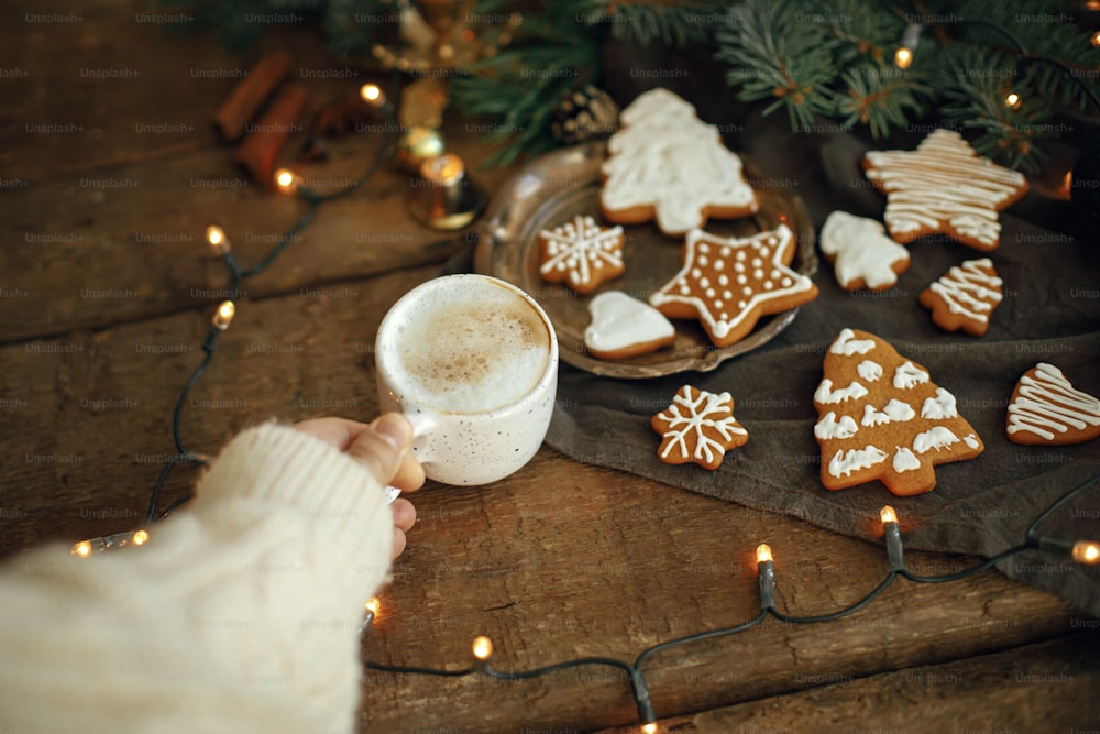 Hand holding warm coffee cup on background of christmas gingerbread cookies, fir branches, warm lights on napkin and rustic wooden table. Hello winter Moody atmospheric image