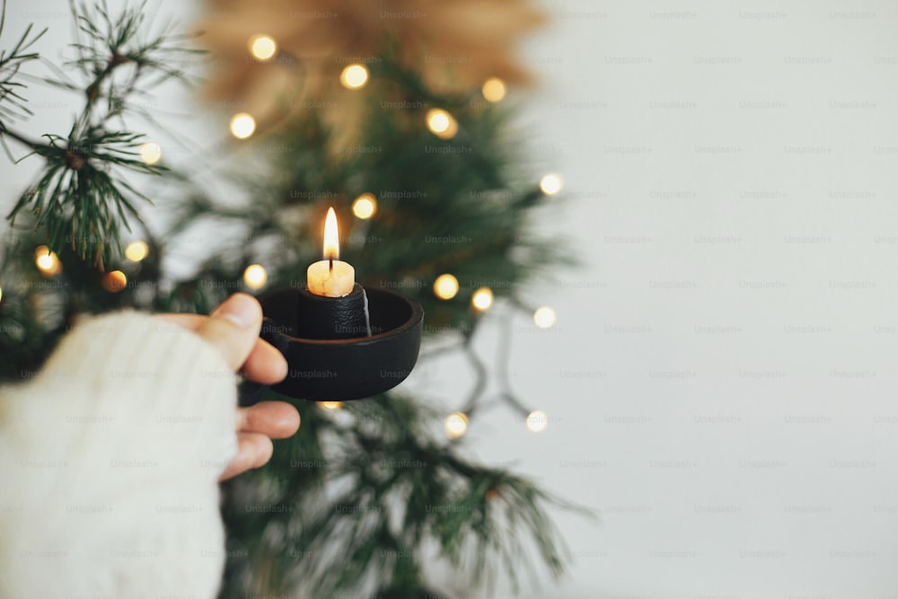 Hand in cozy sweater holding vintage candlestick with burning candle on background of warm lights, fir branches, sweden star in festive scandinavian room. Atmospheric hygge winter home