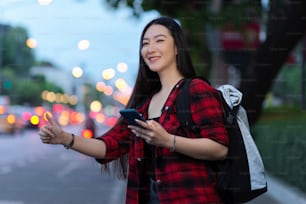 Attraente viaggiatore solista asiatico che chiama il trasporto taxi locale, tenendo smartphone e zaino, luci della città bokeh sullo sfondo