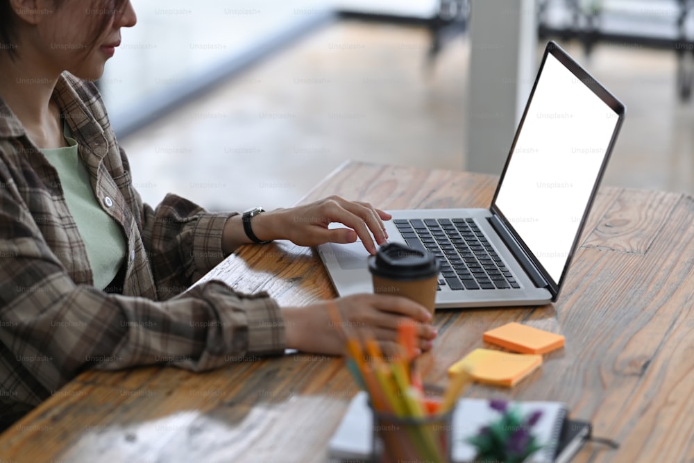 Female freelancer holding coffee cup and working with computer laptop on wooden table.