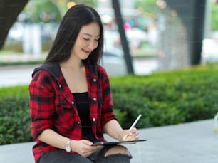 Happy hipster artist woman relaxing in park, using digital tablet and stylus pen to sketching cityscape view and finding inspiration for her digital artwork