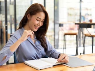 Attractive businesswoman reading list on planner book, blank notebook and tablet on desk, holding glasses, office background