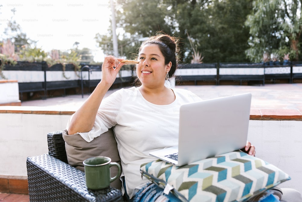 Junge hispanische Frau mit Computer sitzt auf der Terrasse und isst einen Snack in Lateinamerika