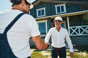 Engineer in white shirt and jeans giving a handshake to builder while staying outside private house