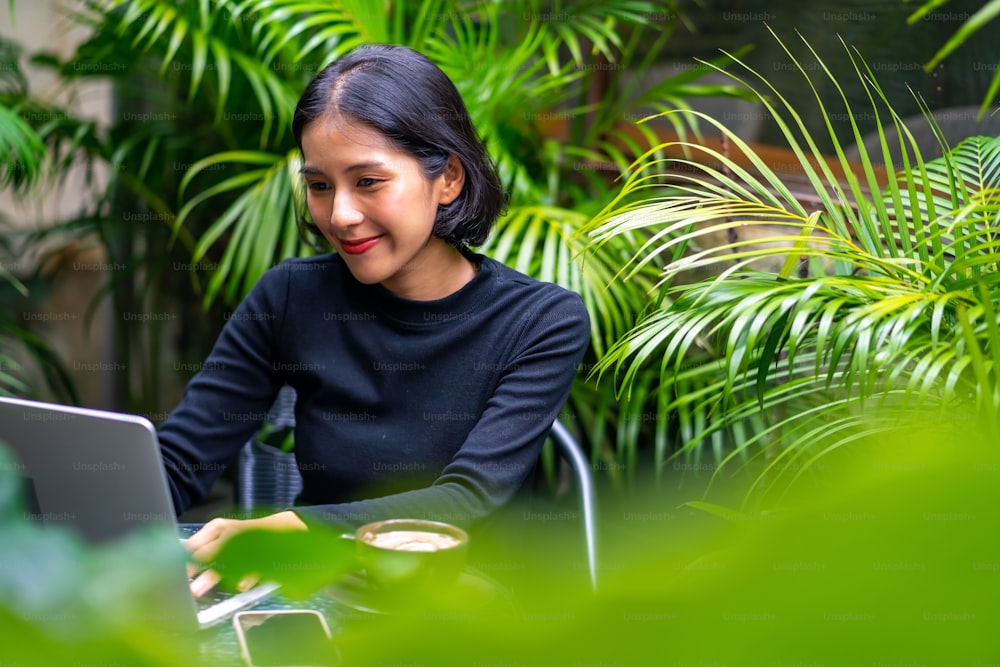Young beautiful Asian woman freelancer working on laptop computer with drinking a cup of coffee in the garden at home. Attractive female using laptop for remote corporate business or online shopping.