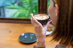 Beautiful Asian woman sitting by the window drinking coffee latte with eating bakery in coffee shop. Beautiful female relax and enjoy weekend activity lifestyle at cafe. Small business food and drink concept.