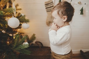 Cute baby girl looking at modern decorated christmas tree with white baubles. Happy stylish toddler little girl standing at xmas tree in festive scandinavian room. Authentic lovely moment