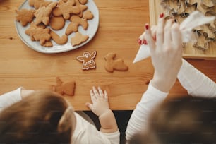 Cute little daughter and mother decorating christmas gingerbread cookies with icing on wooden table.Top view.  Family time, xmas holiday preparations. Mommy daughter authentic lovely moments