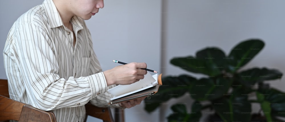 Young man holding pen and making notes on notebook while sitting in home office.