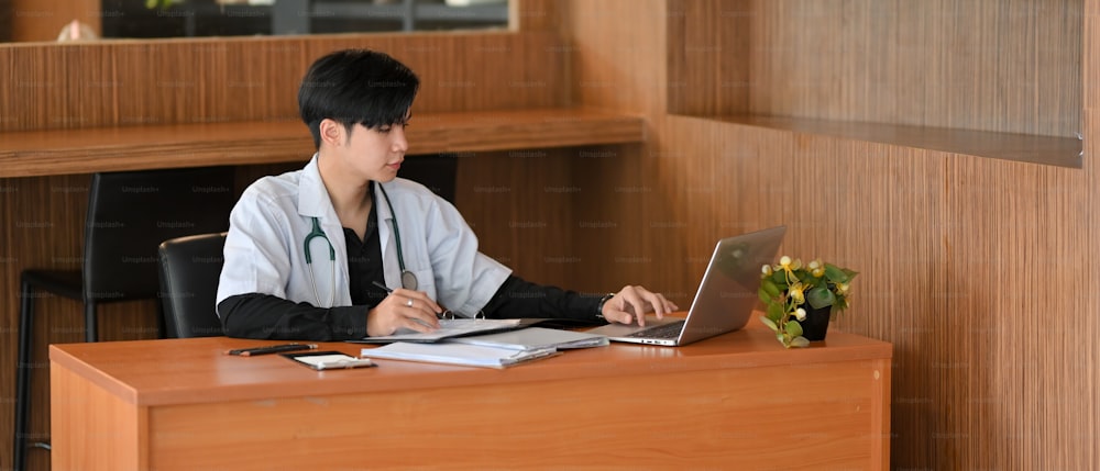 An Asian doctor sits at his desk works on laptop computer, examining the patient's medical history in preparation for an examination and diagnosis.