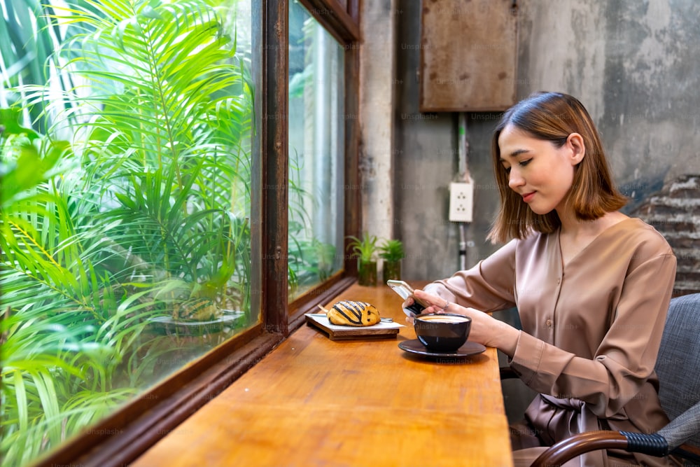 Beautiful Asian woman sitting by the window drinking coffee latte with using smartphone at coffee shop. Beautiful female relax and enjoy weekend activity lifestyle at cafe. Small business food and drink concept