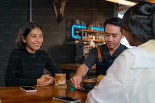 Asian man coffee shop waiter barista serving coffee latte and bakery to woman customer on the table at cafe. Attractive female friend meeting and talking together at restaurant. Small business concept.