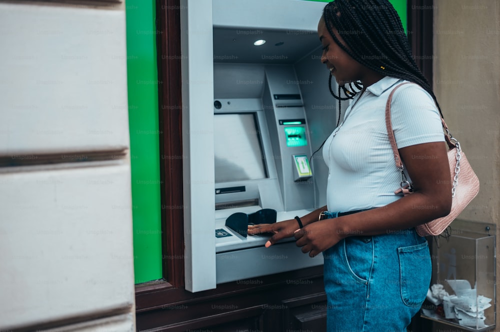 Mulher afro-americana nova que usa o cartão de crédito e a máquina do caixa eletrônico ao retirar o dinheiro
