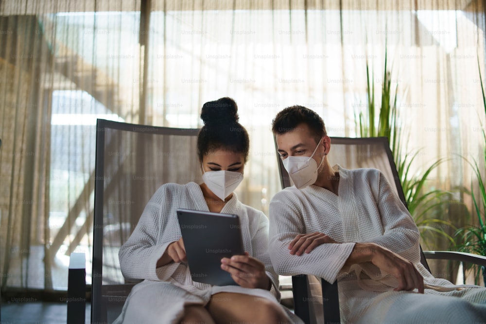 A portrait of young couple with tablet sitting and relaxing in spa resort, coronavirus concept.