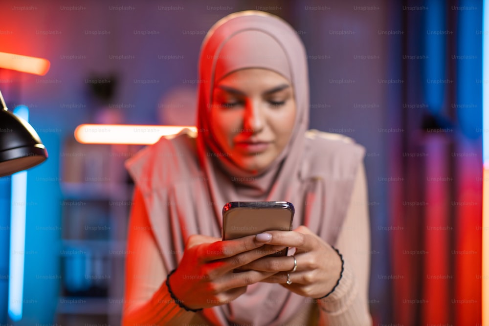 ヒジャーブをかぶった若い女性が机に座り、最新のスマートフォンを手にしています。自宅で夕方に携帯電話でインターネットをサーフィンするイスラム教徒の女性。