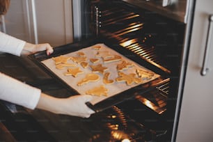 Femme mettant un plateau avec des biscuits de Noël dans un four en gros plan dans une cuisine moderne. Cuisson de biscuits au pain d’épice. Préparation des vacances en famille, culinaire de Noël. Recette de biscuits