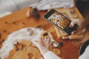 Mujer sosteniendo el teléfono y tomando una foto de galletas de jengibre galletas de navidad en una mesa desordenada plana. Foto de Instagram, redes sociales y blog. Preparativos para las fiestas