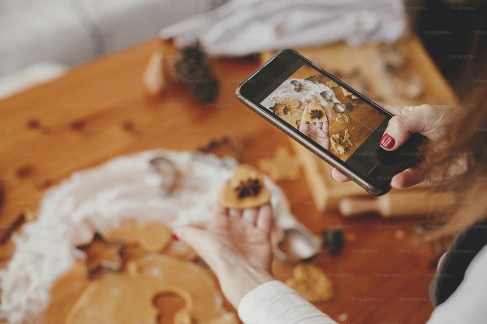 携帯電話を持ち、ジンジャーブレッドクッキーの写真を撮る女性、乱雑なテーブルの上のクリスマスクッキー、平らな横置き。インスタグラムの写真、ソーシャルメディア、ブログ。休日の準備