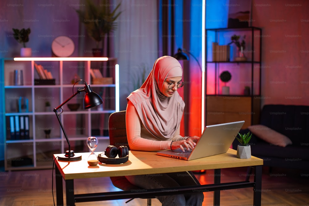 Young woman in hijab and eyeglasses working on modern laptop while sitting at table during evening time. Remote work at home. Freelance and people concept.