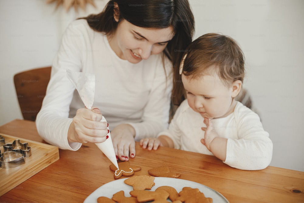 Süße kleine Tochter und Mutter dekorieren Weihnachtslebkuchenplätzchen mit Zuckerguss auf Holztisch in modernem Zimmer. Frohe Familienzeit, Urlaubsvorbereitungen. Frohe Feiertage!