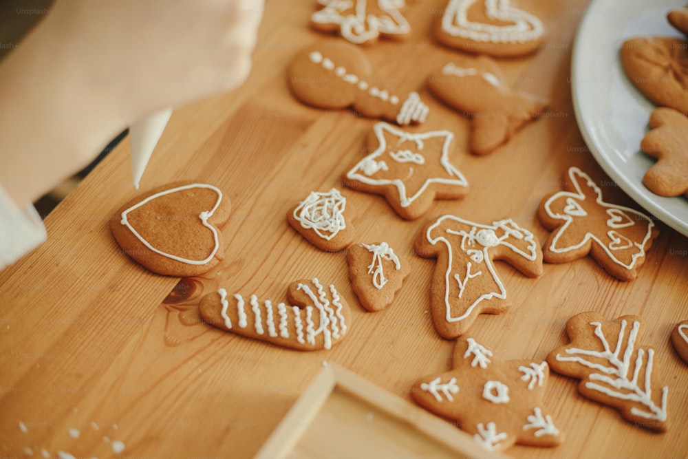 Decorating christmas gingerbread cookies with icing on wooden table close up. Hands decorating baked christmas cookies with sugar frosting. Xmas holiday preparations, atmospheric time