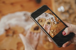 Woman making photo of gingerbread cookies for social media and blog. Hand holding phone and taking photo of heart shaped christmas cookies on messy table flat lay. Holiday preparations
