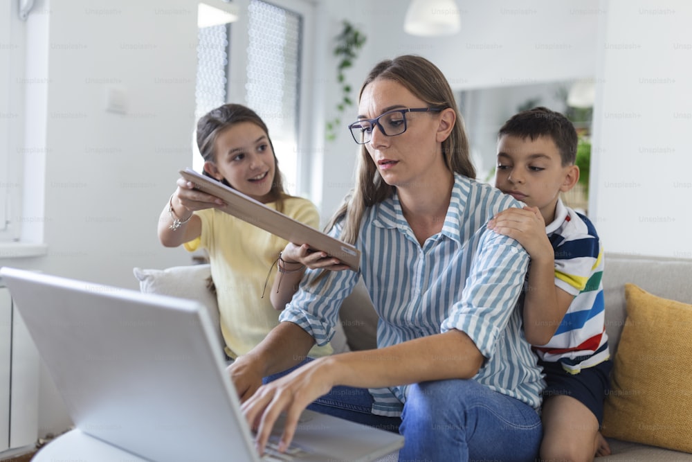 Stressed woman with kids working from home.Tired young mother sitting on sofa and working with laptop and documents while little kids having fun and making noise