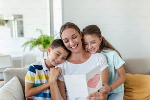 Joyful young mother and little children sitting on sofa and embracing while reading wishes and congratulations with holiday in presented postcard at home