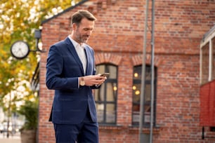 Photo of handsome smiling businessman using mobile phone outdoor on the city street. Thoughtful businessman in elegant suit talking by smartphone.