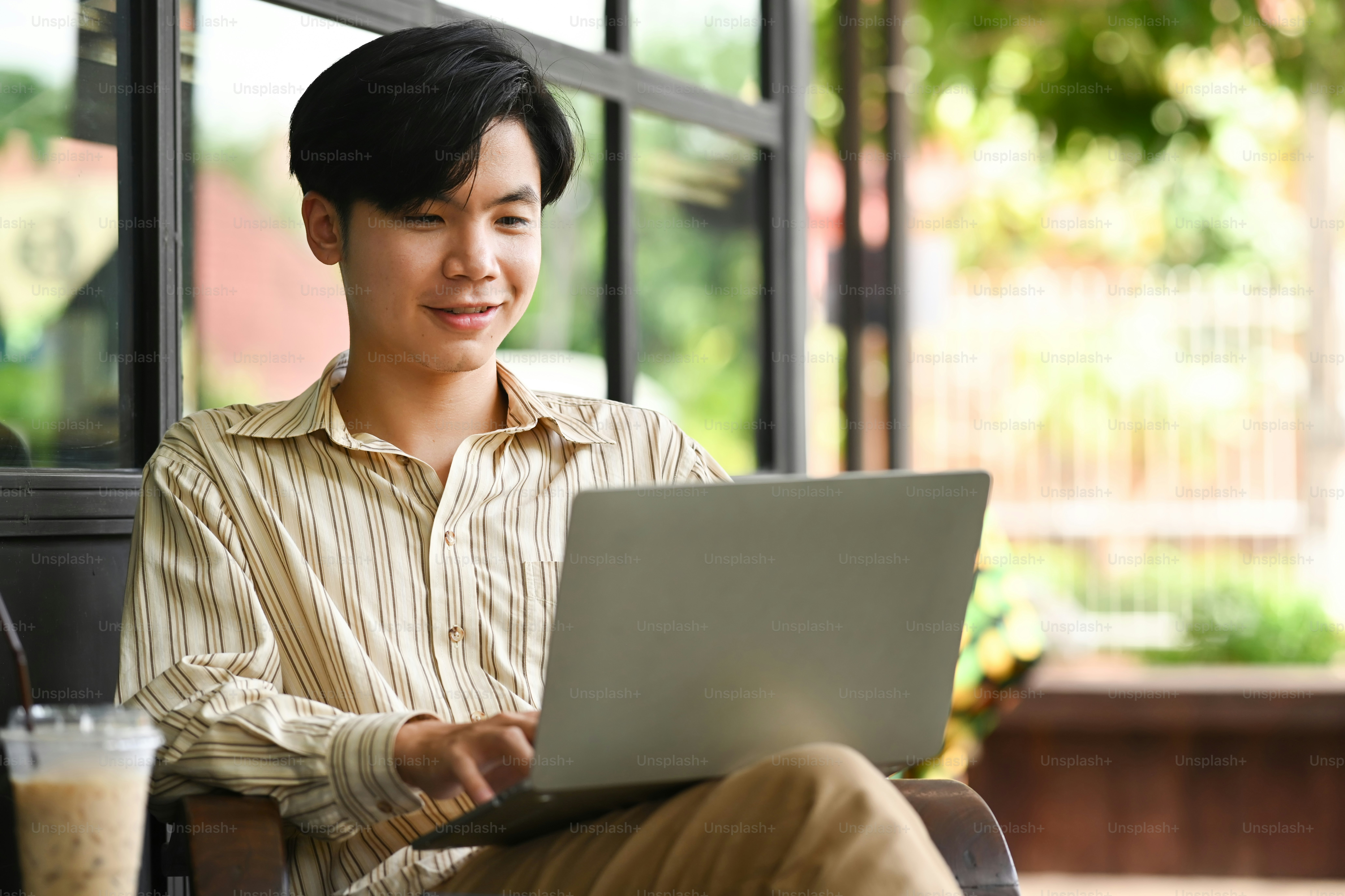 Smiling young Asian man sitting at outdoor cafe and using computer ...