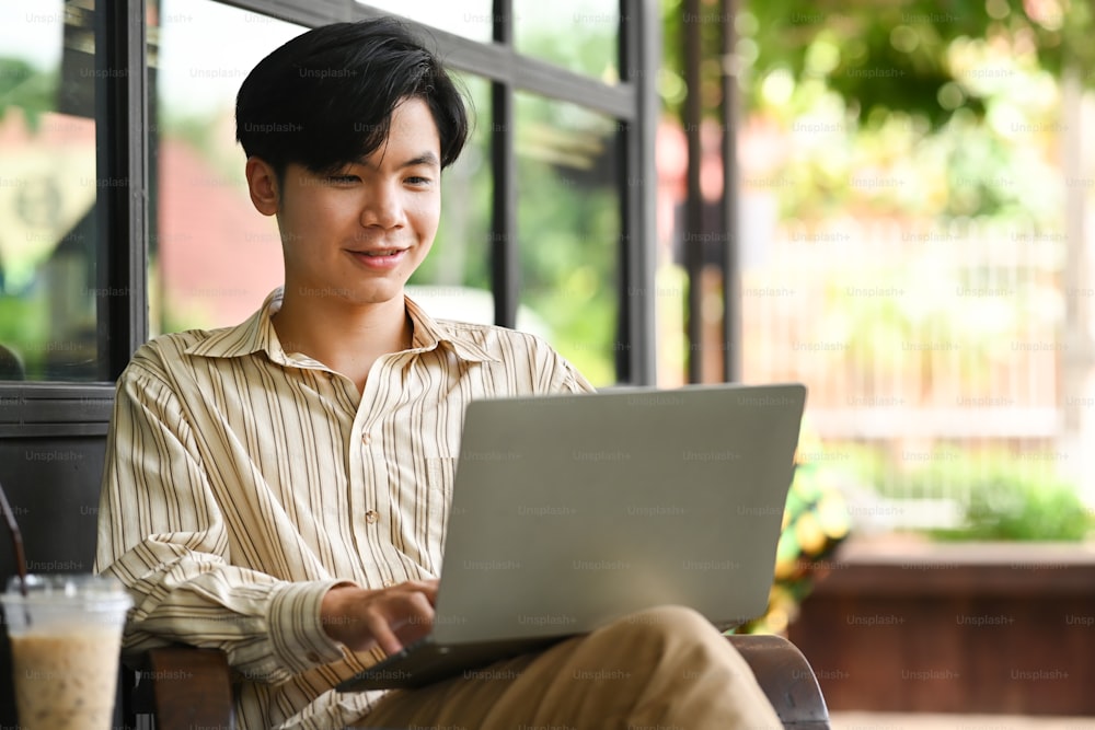 웃고 있는 젊은 아시아 남자가 야외 카페에 앉아 컴퓨터 노트북을 사용하고 있다.