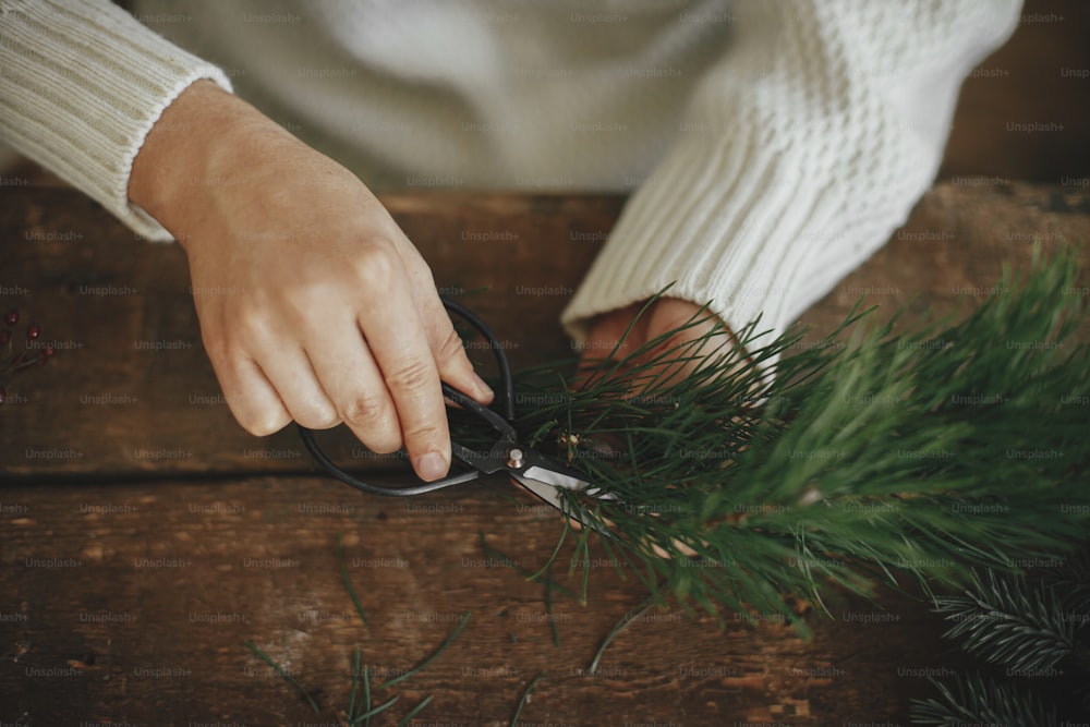 크리스마스 화환 만들기. 아늑한 스웨터를 입은 여자가 소박한 나무 배경에 가위로 소나무 가지를 자르고 손을 닫습니다. 겨울 휴가 준비. 축제 워크샵, 수제 장식