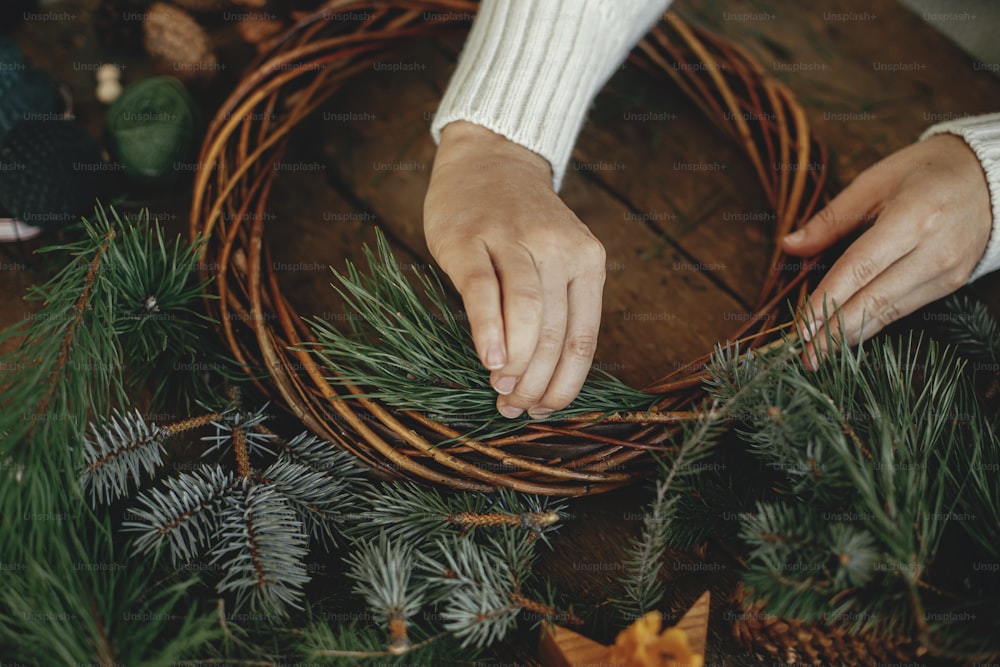 Making christmas wreath. Woman in cozy sweater holding pine branches and arranging christmas wreath on rustic wooden background with pine cones, scissors, thread. Winter holiday preparations