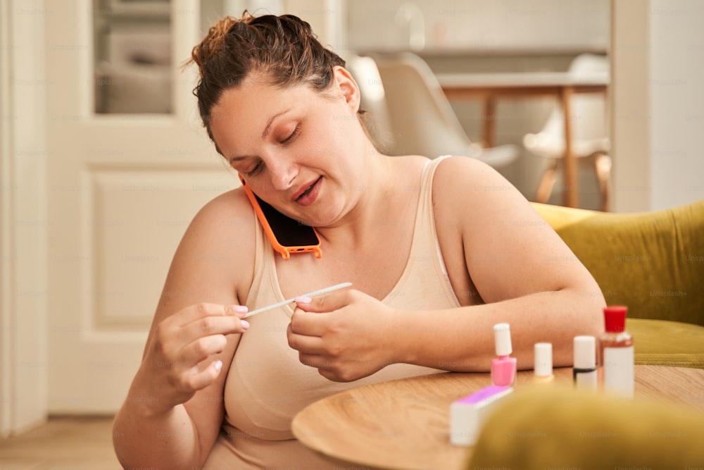 Manicura. Simpática mujer con cuerpo gordo charlando en el teléfono inteligente y preparando manicura con placer. Cuidado de uñas en casa, herramientas de manicura, belleza, arte de uñas, concepto de glamour