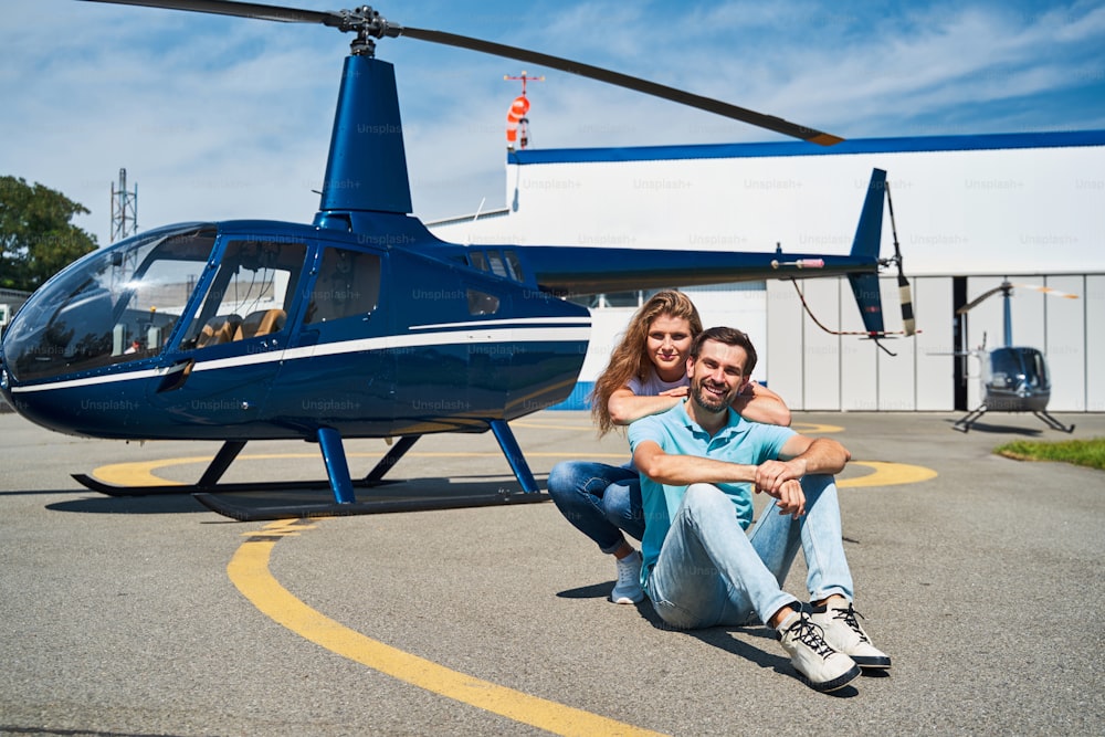Pleased female sitting on her haunches behind happy Caucasian man relaxing on helipad