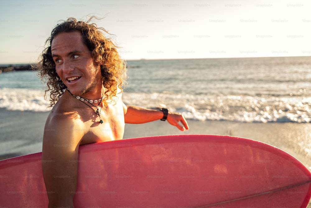 Happy surfer man carrying surfboard to the water on sandy beach. Holiday. Sunset light. Real people lifestyle and water sport concept.