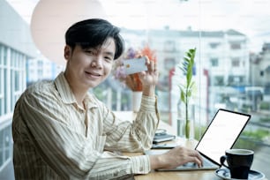 Cheerful young Asian man holding credit card and smiling to camera.
