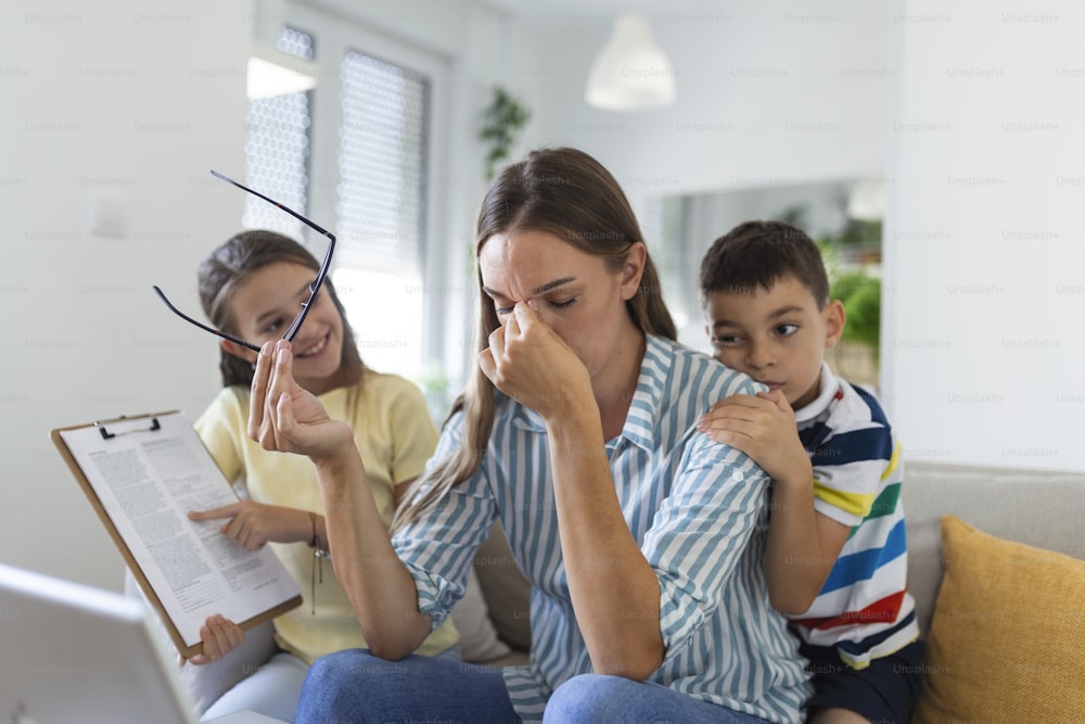 Upset mother having problem with noisy naughty daughter and son jumping on couch and screaming, demanding attention, frustrated mum tired of difficult child trying to work from home