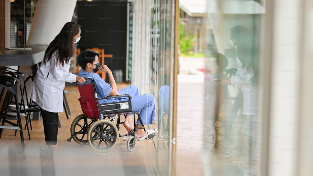 Female doctor and male patient in wheelchair wear face mask look out the window in hospital corridor