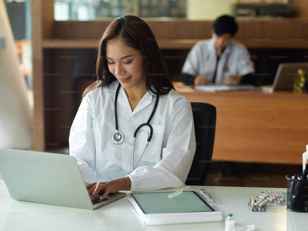 Asian young female doctor in white gown with stethoscope working on laptop computer in office