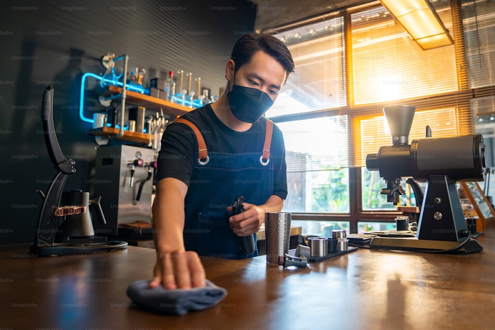 Asian man waiter barista wearing protective face mask during COVID-19 pandemic using towel sweeping counter bar with alcohol sanitizer. Male coffee shop owner cleaning up coffee shop for service to customer. Small business concept.
