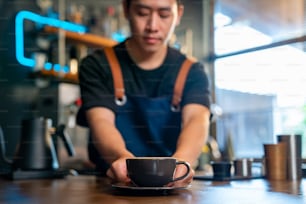 Asiatischer Mann Barista macht heißen Kaffee Latte in Kaffeetasse für Kunden an der Bartheke im Café. Männlicher Coffeeshop-Kellner, der dem Kunden heißen Kaffee mit Milch serviert. Konzept des Restaurantbesitzers für kleine Unternehmen.