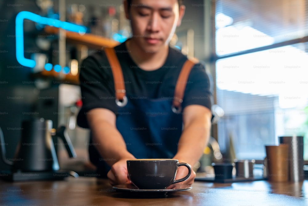 カフェのバーカウンターで、コーヒーカップでホットコーヒーラテを作るアジア人男性のバリスタ。ミルク入りのホットコーヒーを客に振る舞う男性喫茶店のウェイター。中小企業のレストランのオーナーのコンセプト。