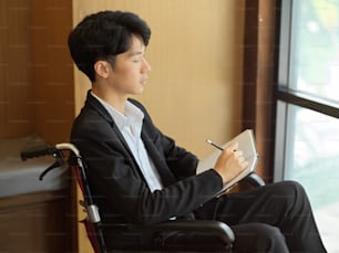 Young businessman in wheelchair against the window, writing ideas on notebook
