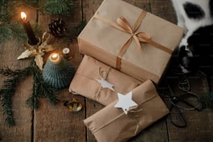 Elegantes regalos de navidad envueltos en papel artesanal, lindo gato, vela, tijeras, ramas de abeto en madera rústica. Presentación moderna simple de Navidad ecológica, imagen atmosférica de mal humor.