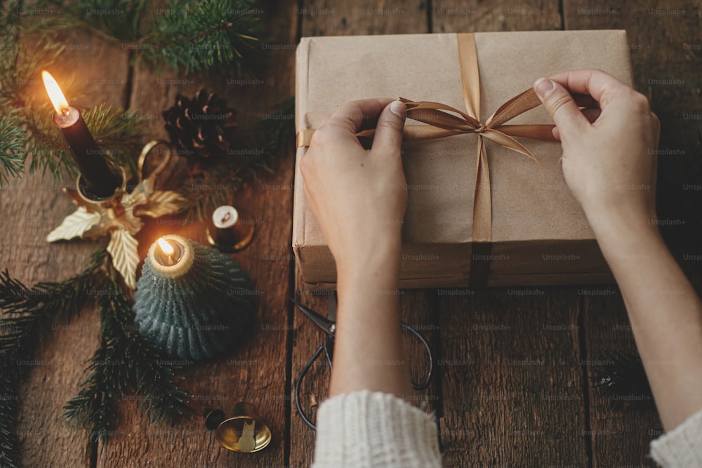 Hände verpacken stilvolles Weihnachtsgeschenk in Bastelpapier auf rustikalem Holzgrund mit Kerze, Schere, Tannenzweigen. Moderne einfache umweltfreundliche Weihnachten präsentieren, skandinavisches atmosphärisches stimmungsvolles Bild