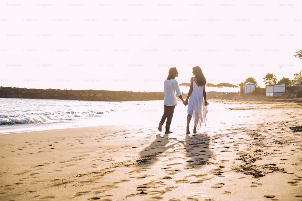 Lindo casal apaixonado caminhando na praia tropical, desfrutando de lua de mel e férias de verão. Mulher e homem de mãos dadas, olhando um para o outro. Estilo de vida de pessoas reais. Hora do pôr-do-sol.