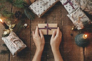 Christmas rustic gift flat lay. Hands holding stylish christmas gift wrapped in craft paper on rustic wooden table with candles, scissors, fir branches. Merry Christmas! Atmospheric moody image